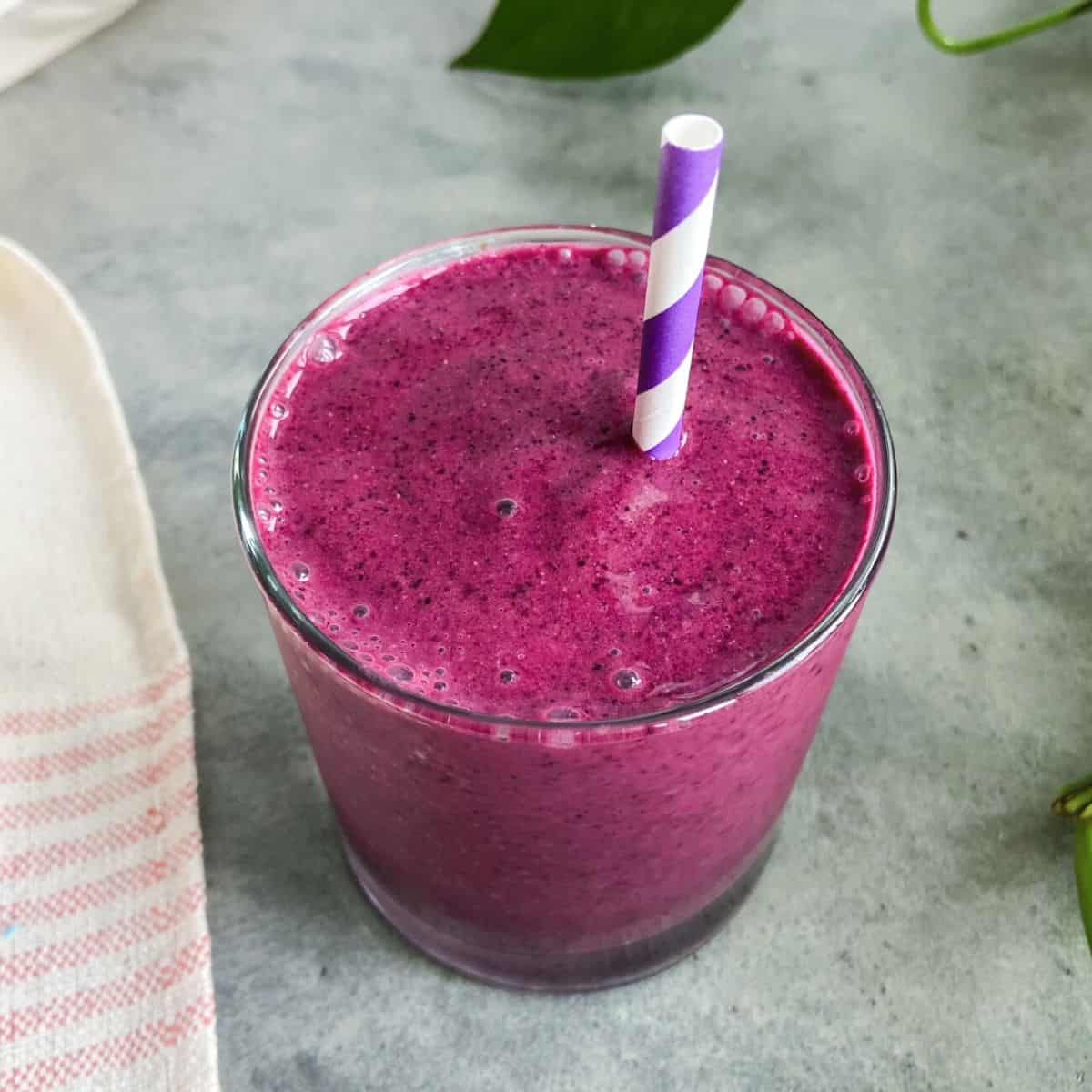A vibrant purple smoothie served in a clear glass with a purple striped straw on a light grey surface.