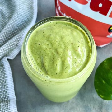 A creamy green smoothie in a glass on a gray surface with a tea towel and red protein powder bag in the background.