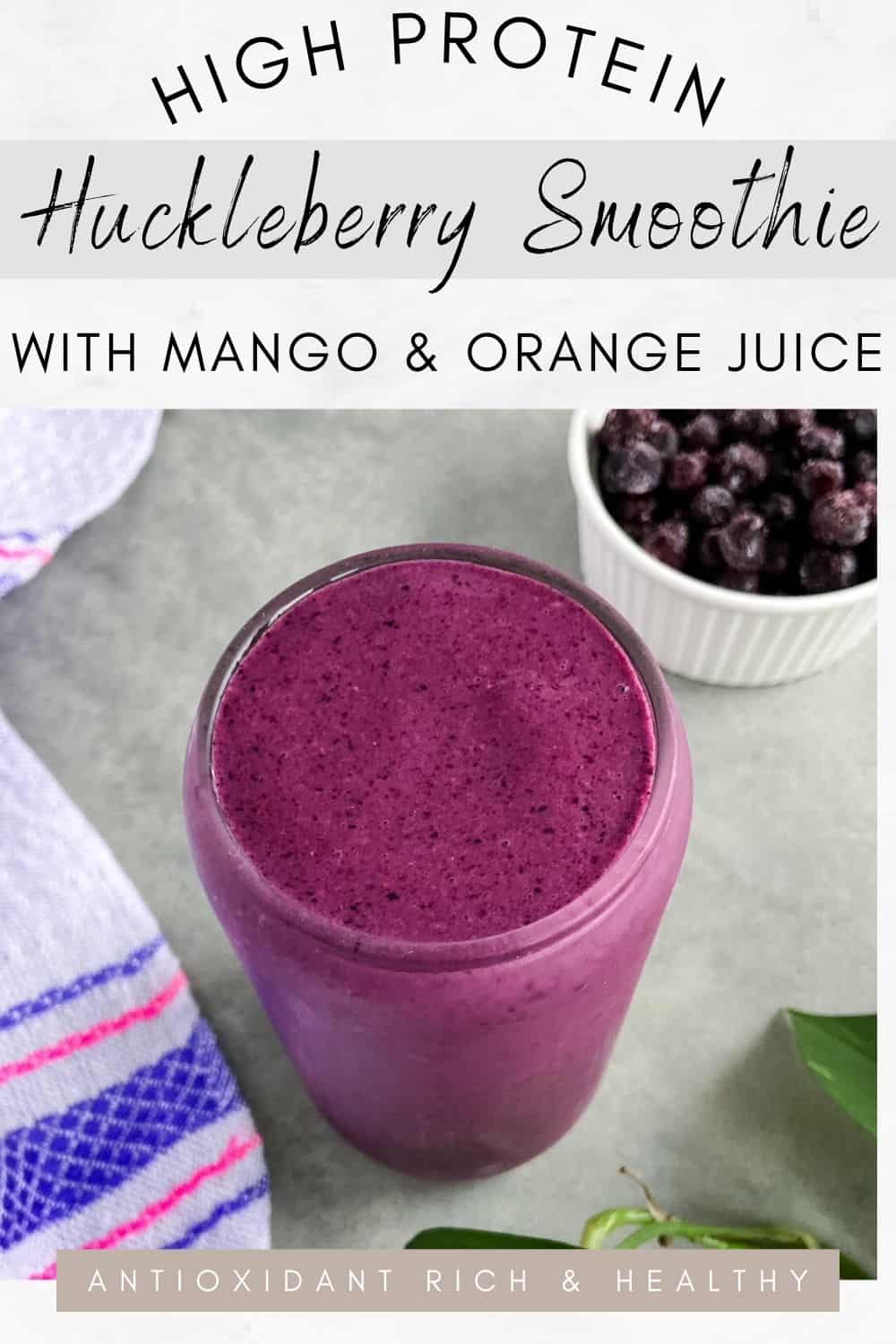 A glass of thick purple berry smoothie on a light countertop with a small bowl of frozen berries huckleberries, a striped kitchen towel, and green leaves nearby.