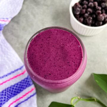 A glass of thick purple huckleberry smoothie on a light countertop, with a small bowl of frozen berries and a striped kitchen towel in the background.