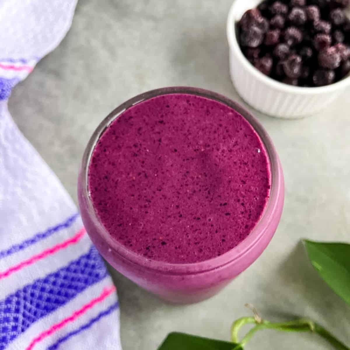 A glass of thick purple huckleberry smoothie on a light countertop, with a small bowl of frozen berries and a striped kitchen towel in the background.