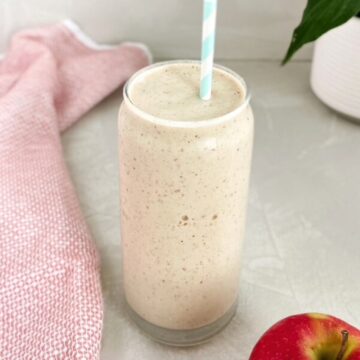 Apple protein smoothie in a clear glass with a straw, creamy lightly slushy texture, and subtle pumpkin pie spice flecks, styled on a light countertop with a tea towel and fresh apple nearby.