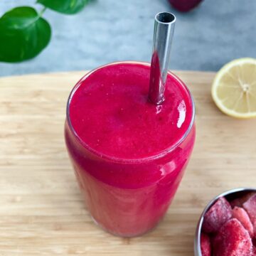 Bright pink smoothie in a clear glass with a metal straw, set on a wooden board with lemon and frozen fruit nearby.