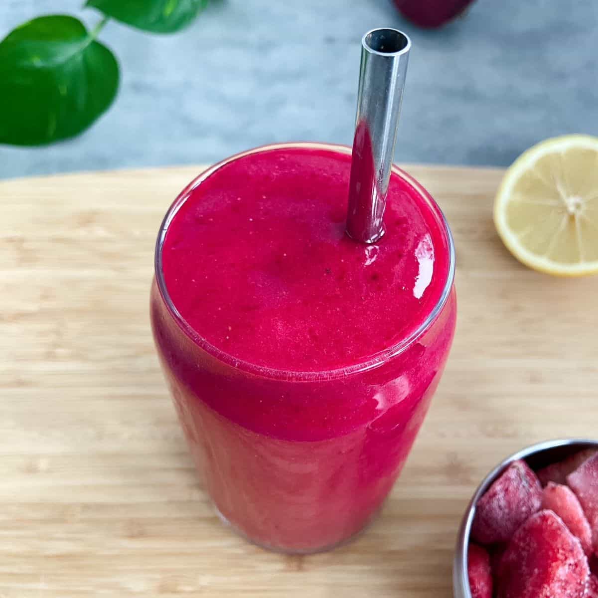Bright pink smoothie in a clear glass with a metal straw, set on a wooden board with lemon and frozen fruit nearby.