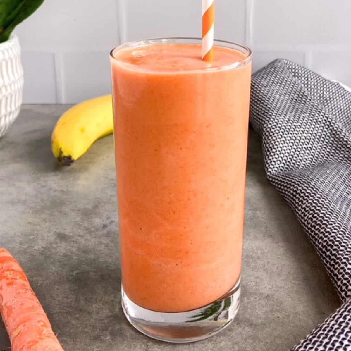 A tall glass of creamy carrot banana smoothie with a striped straw on a gray surface, with a carrot and banana in the background.