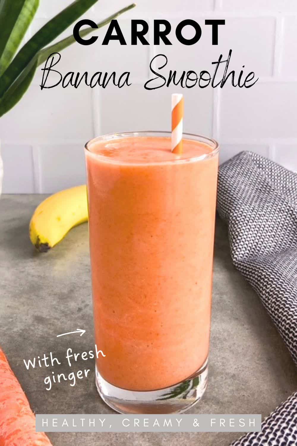 A tall glass of creamy carrot banana smoothie with a striped straw on a gray surface, with a carrot, tea towel and banana in the background.