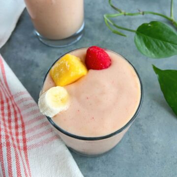 Creamy mango strawberry banana smoothie in a glass topped with a frozen mango cube, a strawberry, and a slice of banana on a gray surface with plant leaves and a tea towel beside the smoothie.