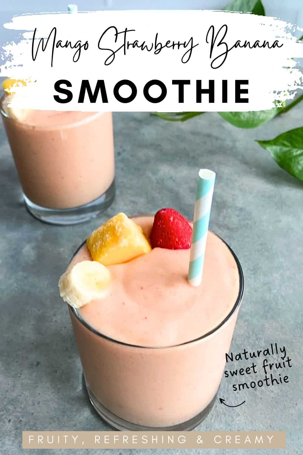 Creamy mango strawberry banana smoothie in a glass topped with a frozen mango cube, a strawberry, and a slice of banana, with a second smoothie in the background on a gray surface.