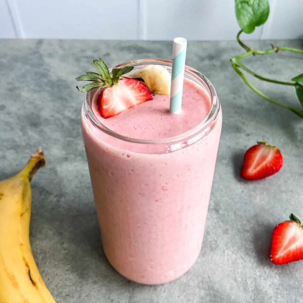 A glass of creamy pink strawberry banana smoothie topped with a fresh strawberry and banana slice, sitting on a grey surface with whole strawberries and a banana in the background.