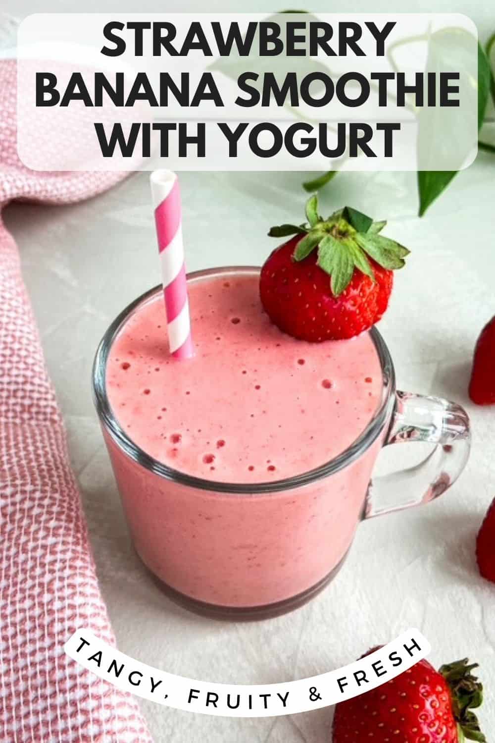 A creamy pink strawberry banana smoothie with yogurt in a clear glass mug, topped with a fresh strawberry and a striped straw, set on a light surface with whole strawberries and a pink cloth nearby.
