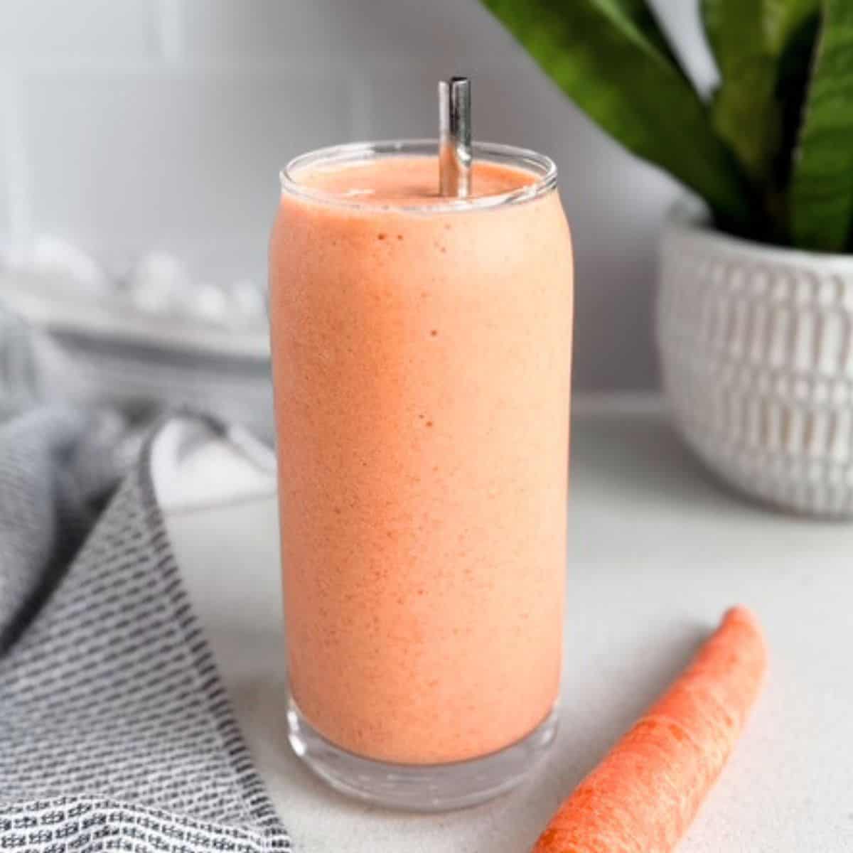 Creamy carrot cake protein smoothie in a tall clear glass with a metal straw and a whole carrot beside it