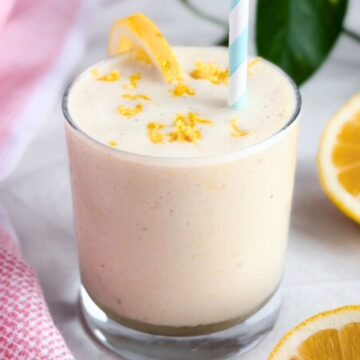 A creamy smoothie in a clear glass topped with fresh lemon zest and a lemon slice, with a striped blue straw and sliced lemons beside and a pink tea towel beside it on a light background.
