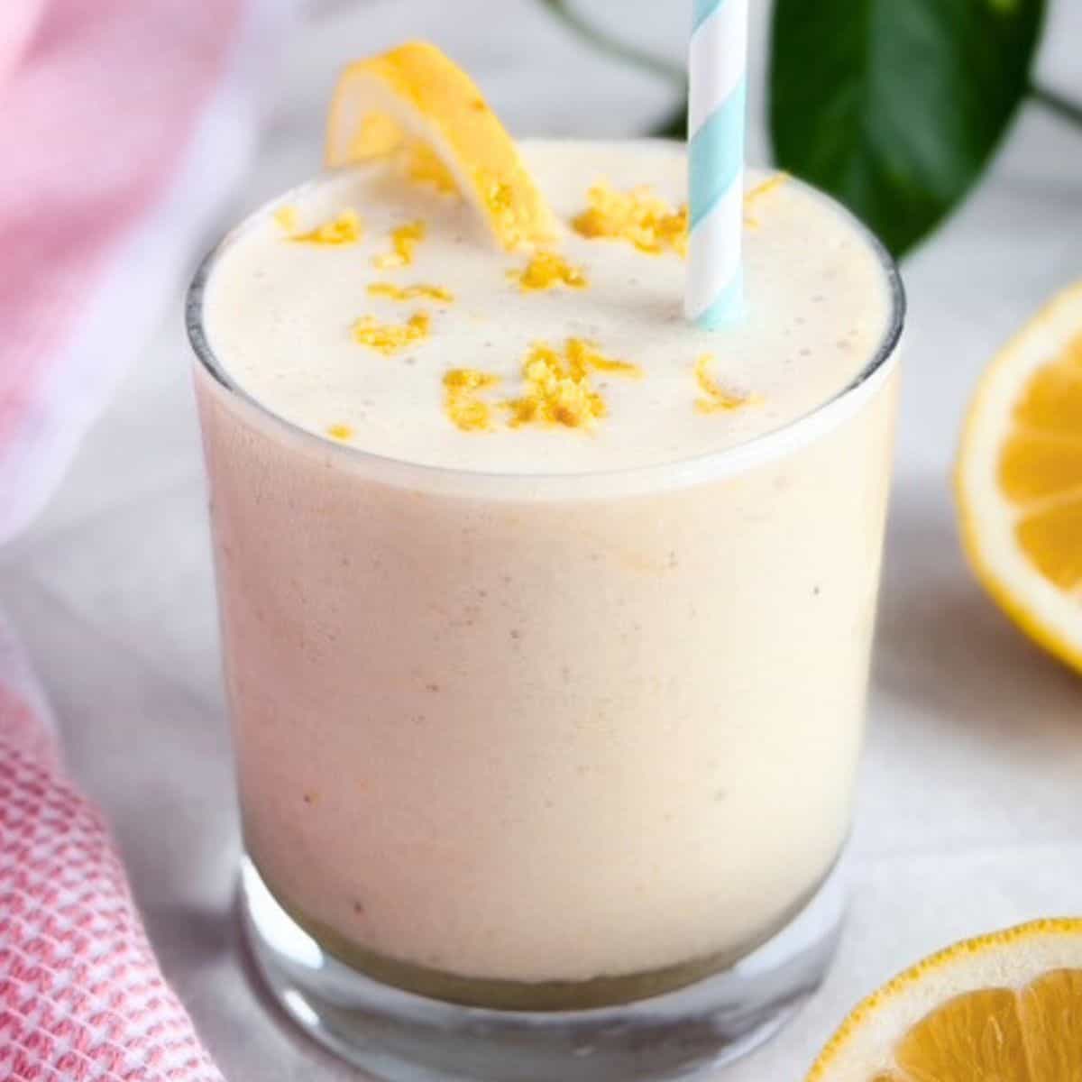 A creamy smoothie in a clear glass topped with fresh lemon zest and a lemon slice, with a striped blue straw and sliced lemons beside and a pink tea towel beside it on a light background.