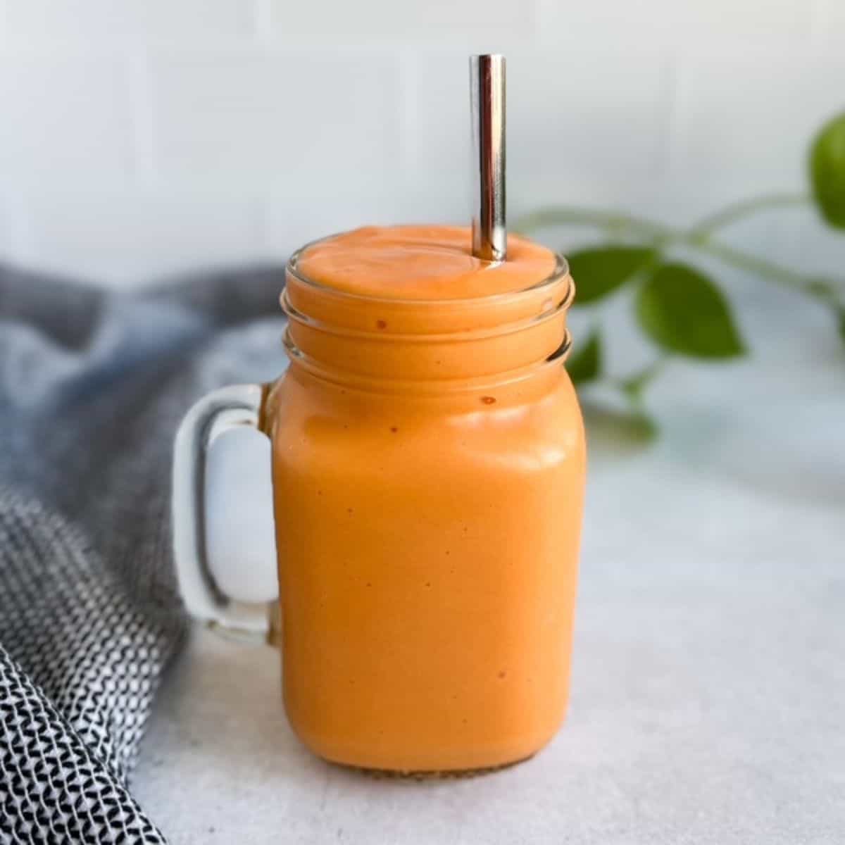 Creamy mango carrot smoothie in a glass jar with a metal straw on a light countertop with a soft neutral background.