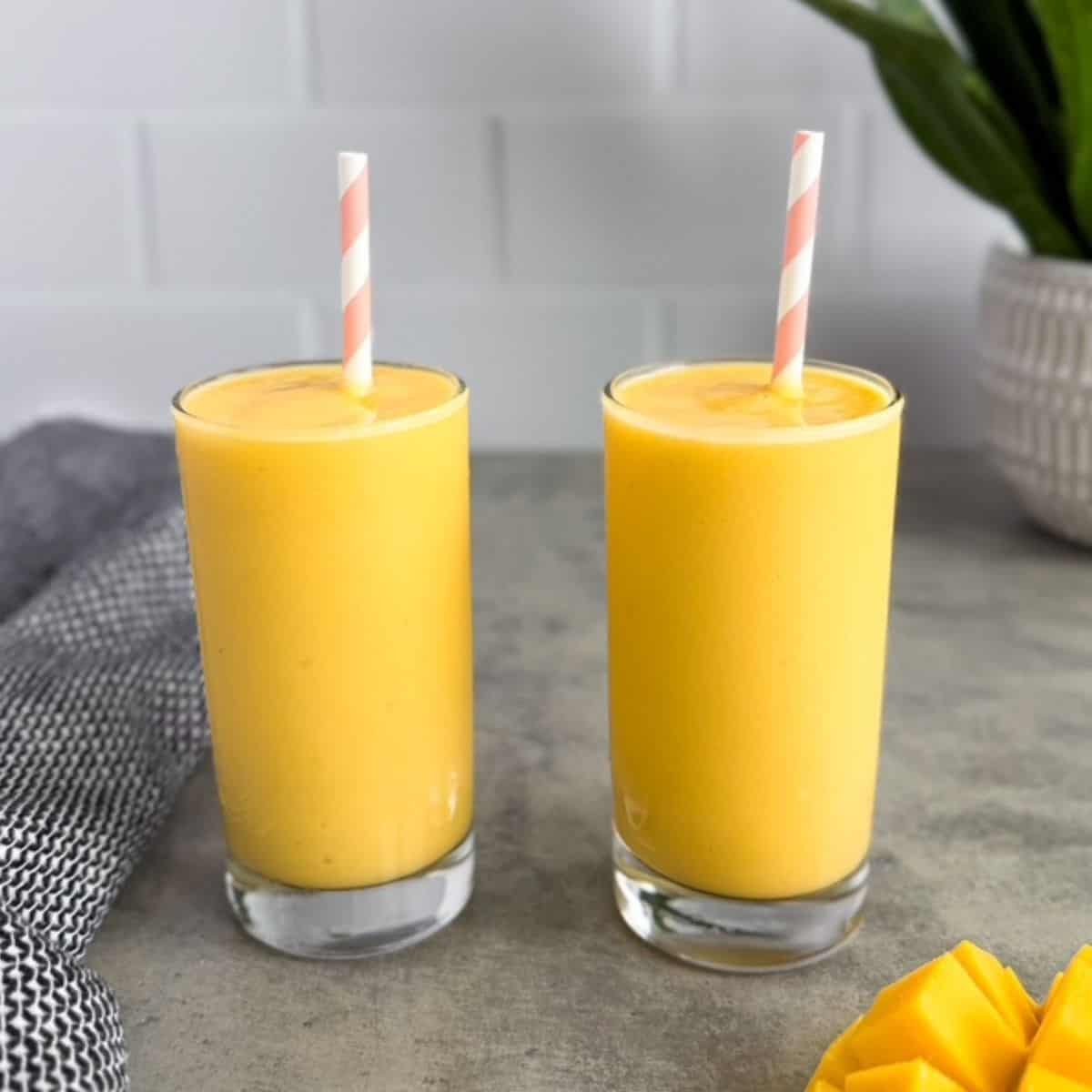 Two glasses of creamy mango coconut smoothie with striped straws on a gray surface with fresh mango pieces nearby