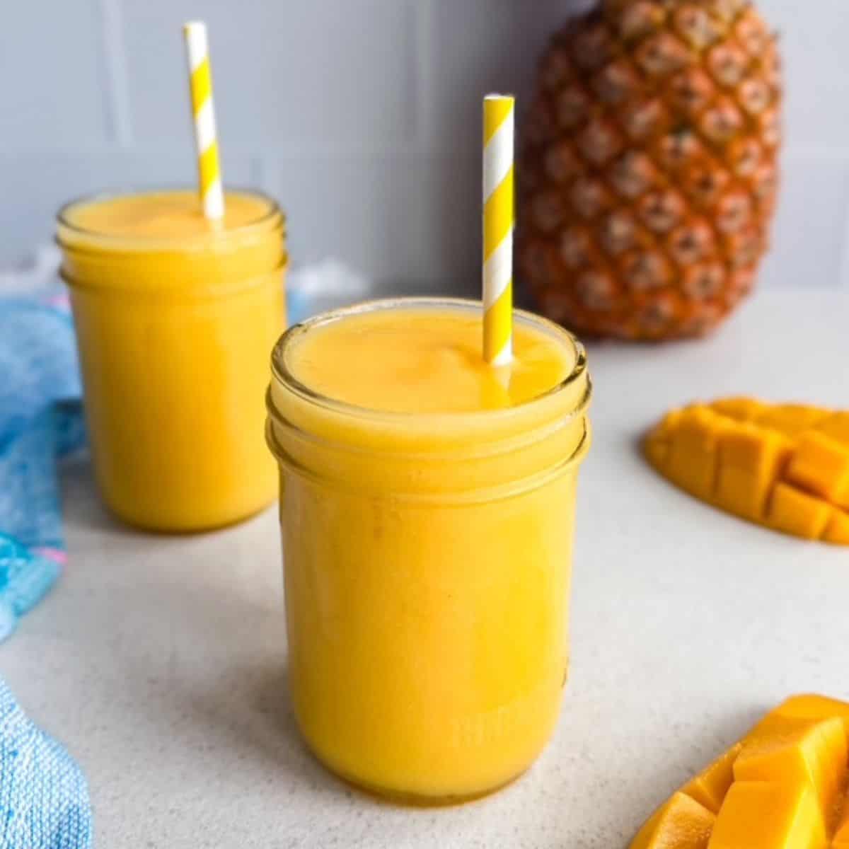 Glass jars of mango pineapple smoothie with striped straws on a countertop with fresh mango slices and a whole pineapple in the background