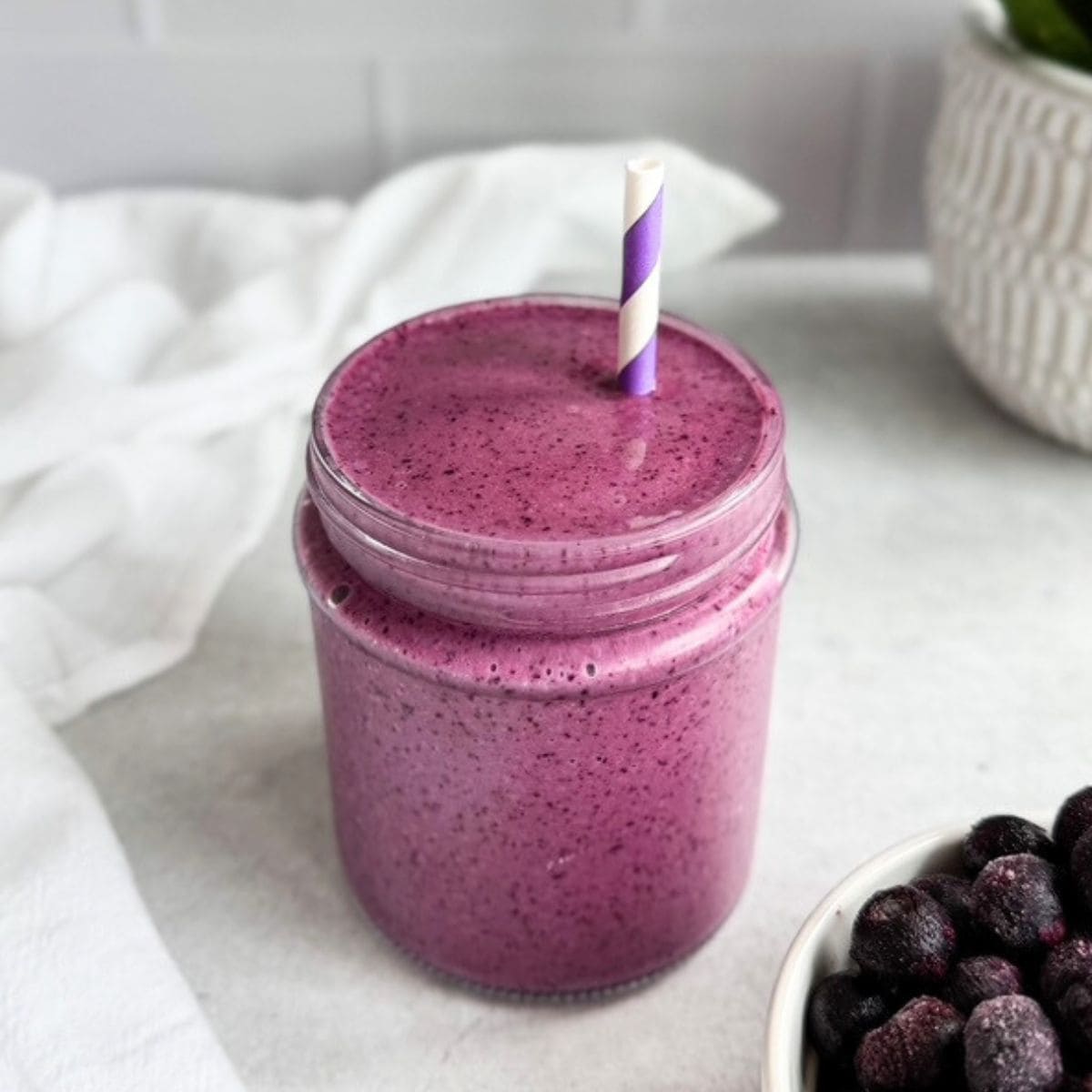Creamy purple blueberry smoothie in a glass jar with a striped straw, with frozen blueberries in a small bowl beside it.