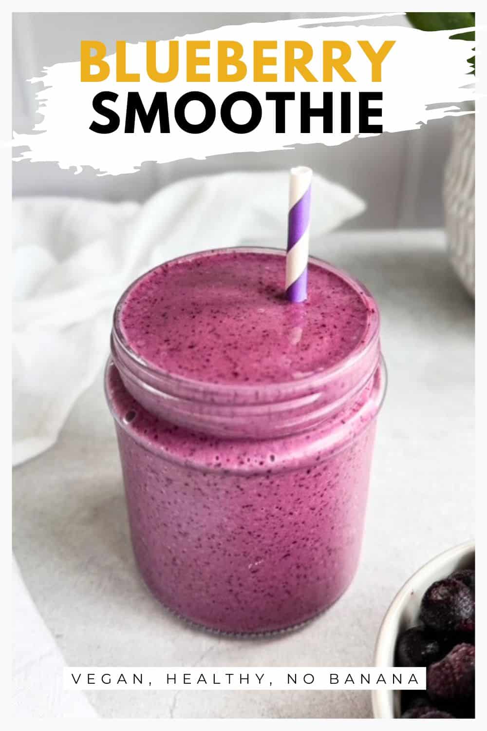 Creamy blueberry smoothie in a glass jar with a purple striped straw, with frozen blueberries in a small bowl on the side.