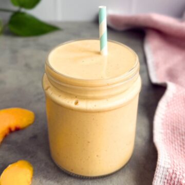 Peach smoothie in a glass jar with a straw on a grey surface, with peach slices and a pink tea towel on the side.