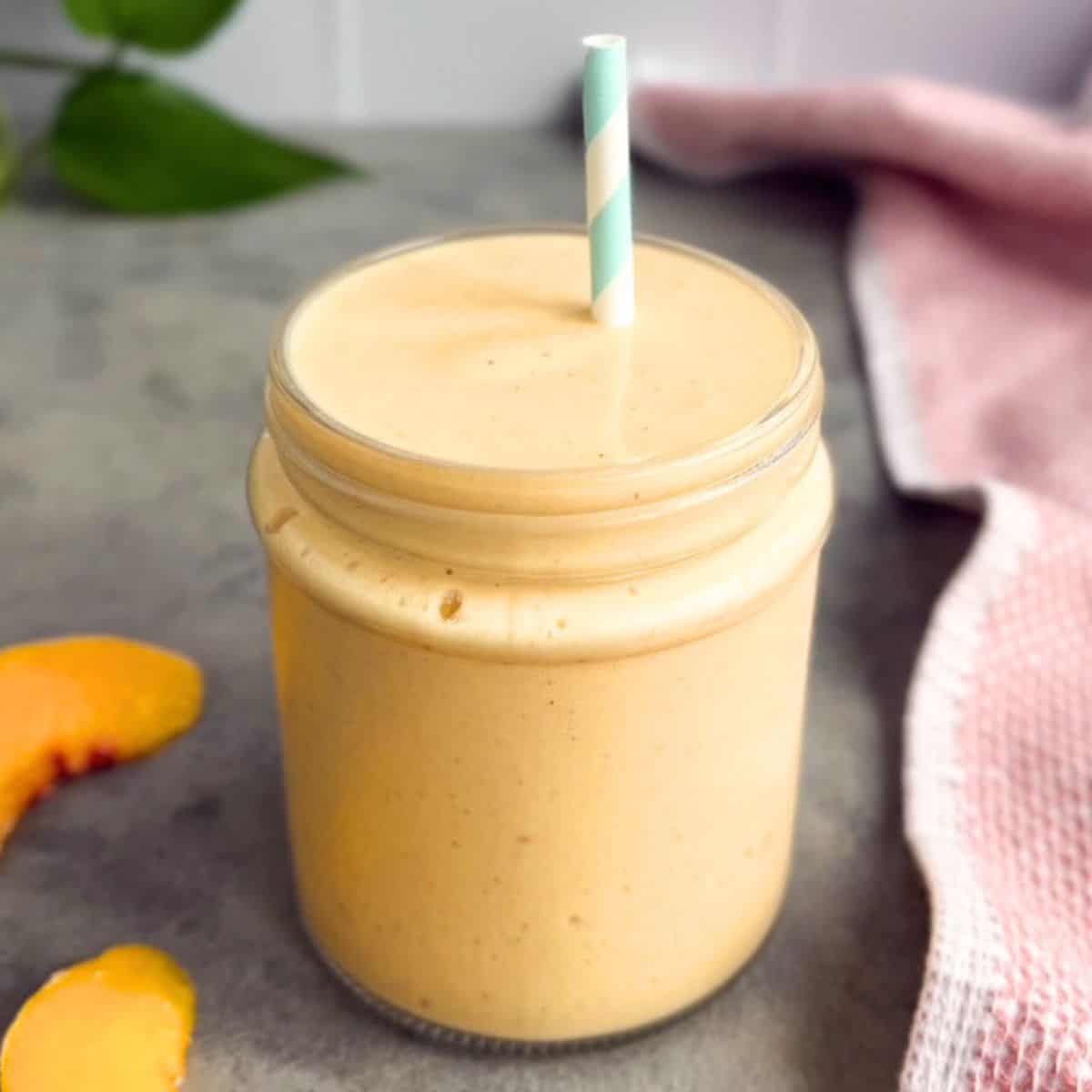 Peach smoothie in a glass jar with a straw on a grey surface, with peach slices and a pink tea towel on the side.