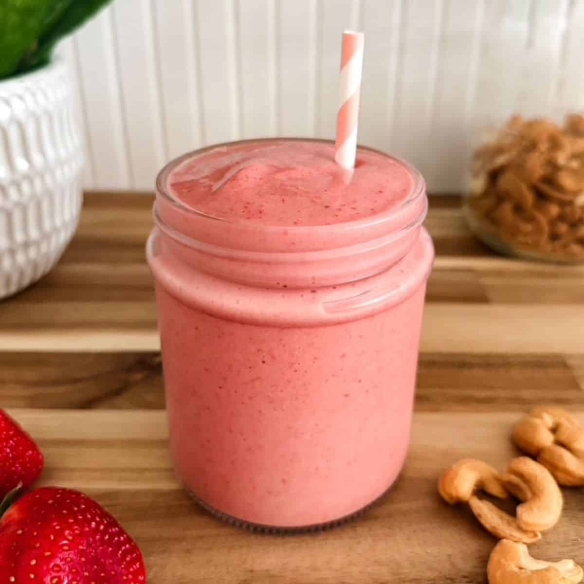 Creamy strawberry cashew smoothie in a glass jar with fresh strawberries and raw cashews on a wooden board.