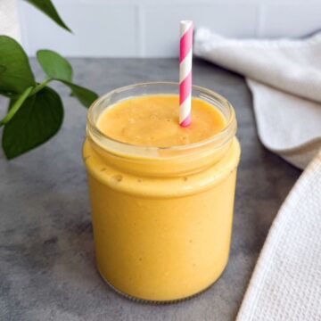Creamy mango smoothie in a glass jar with a pink striped straw on a grey countertop with a plant and tea towel on the side.