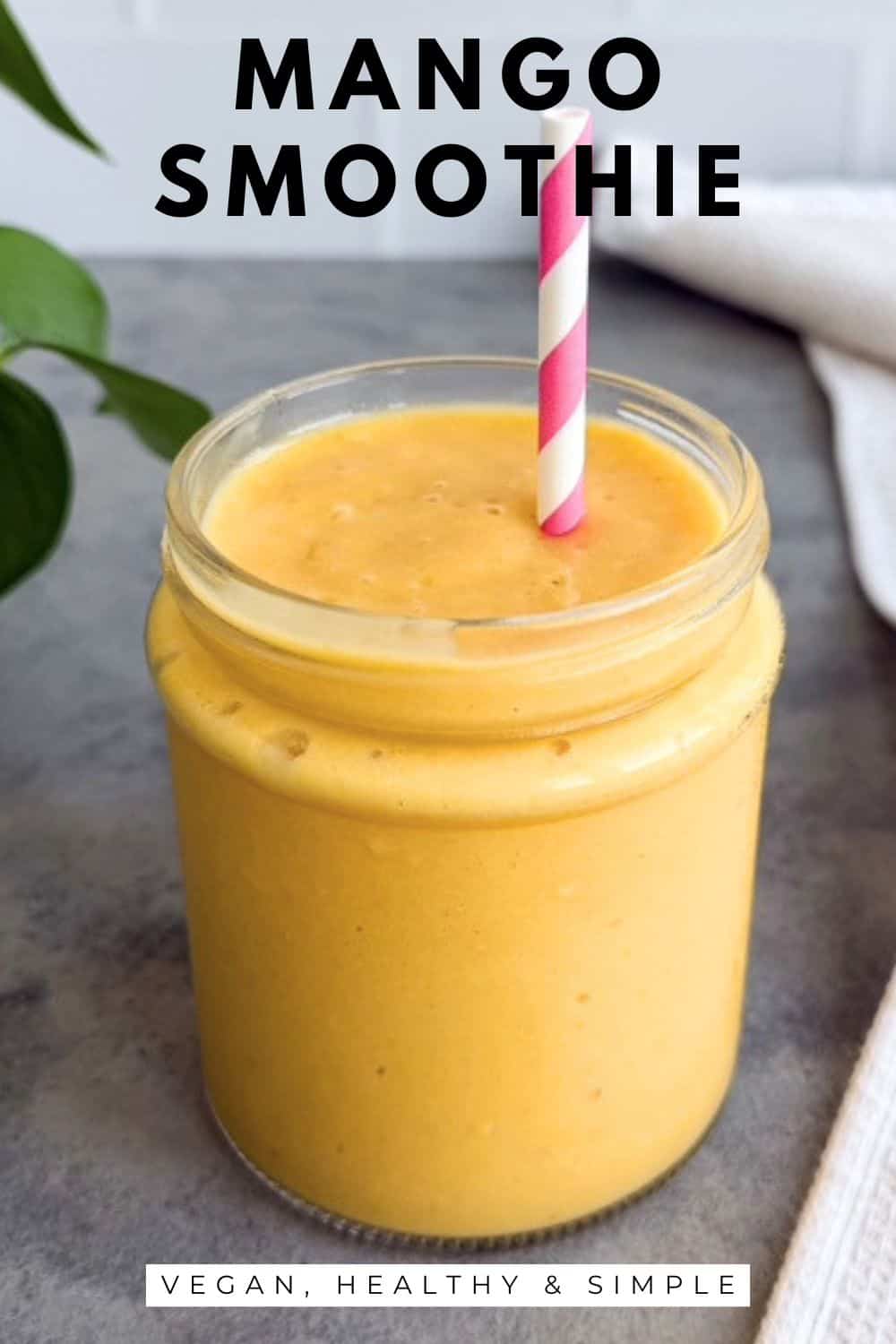Creamy mango smoothie in a glass jar with a pink striped straw on a grey countertop with a plant and tea towel on the side.