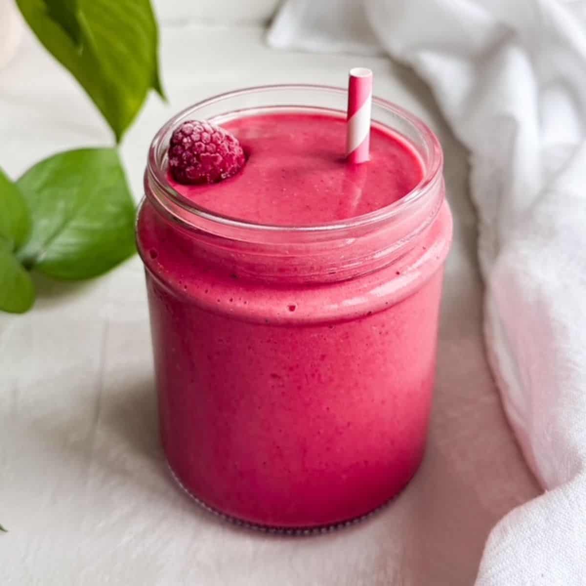 A glass jar filled with a bright pink raspberry smoothie topped with a frozen raspberry and a striped straw, with a white tea towel and green plant leaves in the background.