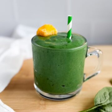 Green smoothie in a glass mug with a mango chunk and striped green straw sitting on a wood counter with a white tea towel laying beside the smoothie glass.