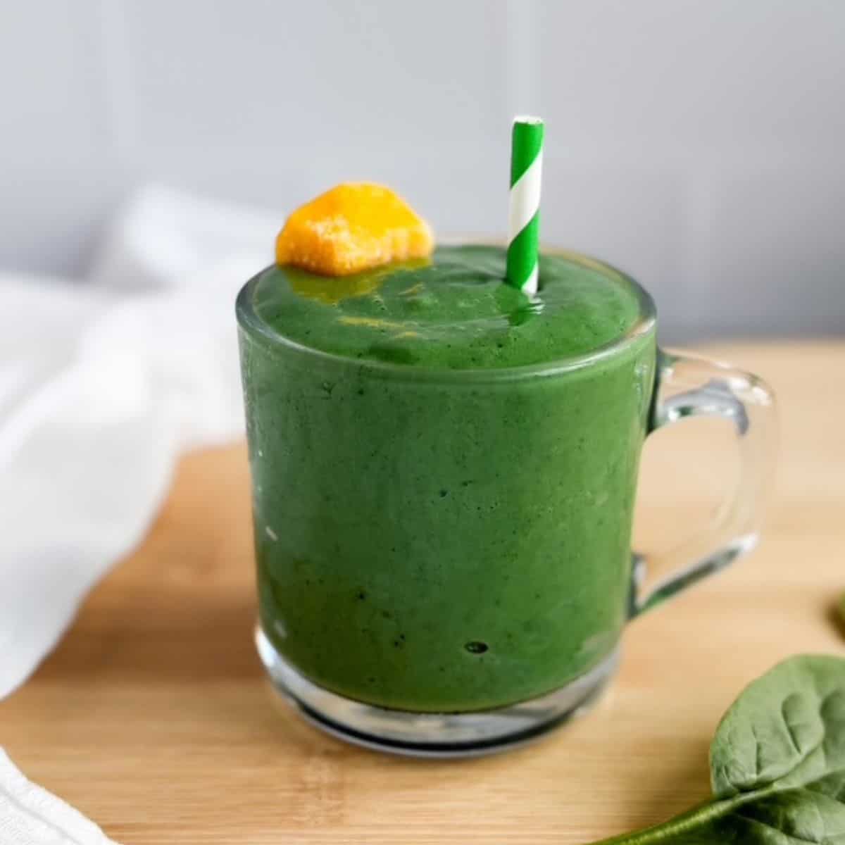 Green smoothie in a glass mug with a mango chunk and striped green straw sitting on a wood counter with a white tea towel laying beside the smoothie glass.
