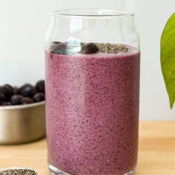 Creamy purple blueberry breakfast smoothie in a glass, topped with whole blueberries and chia seeds, with fresh blueberries in the background.