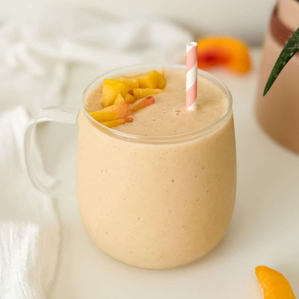 Creamy peach yogurt smoothie topped with peach pieces in a glass with a pink striped straw.