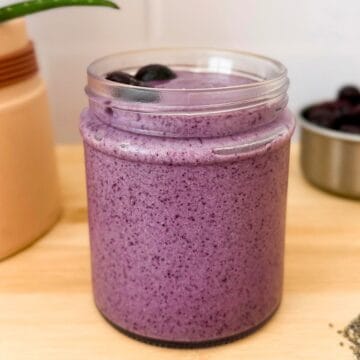 Blueberry smoothie in a clear glass cup topped with half-frozen blueberries.