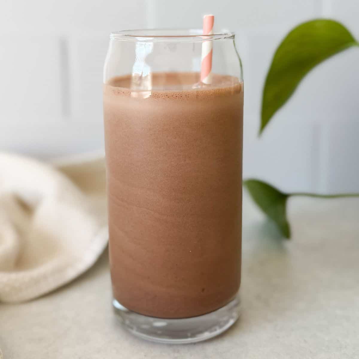 A chocolate protein shake in a tall clear glass with a pink and white striped straw, sitting on a light countertop with green plant leaves in the background.